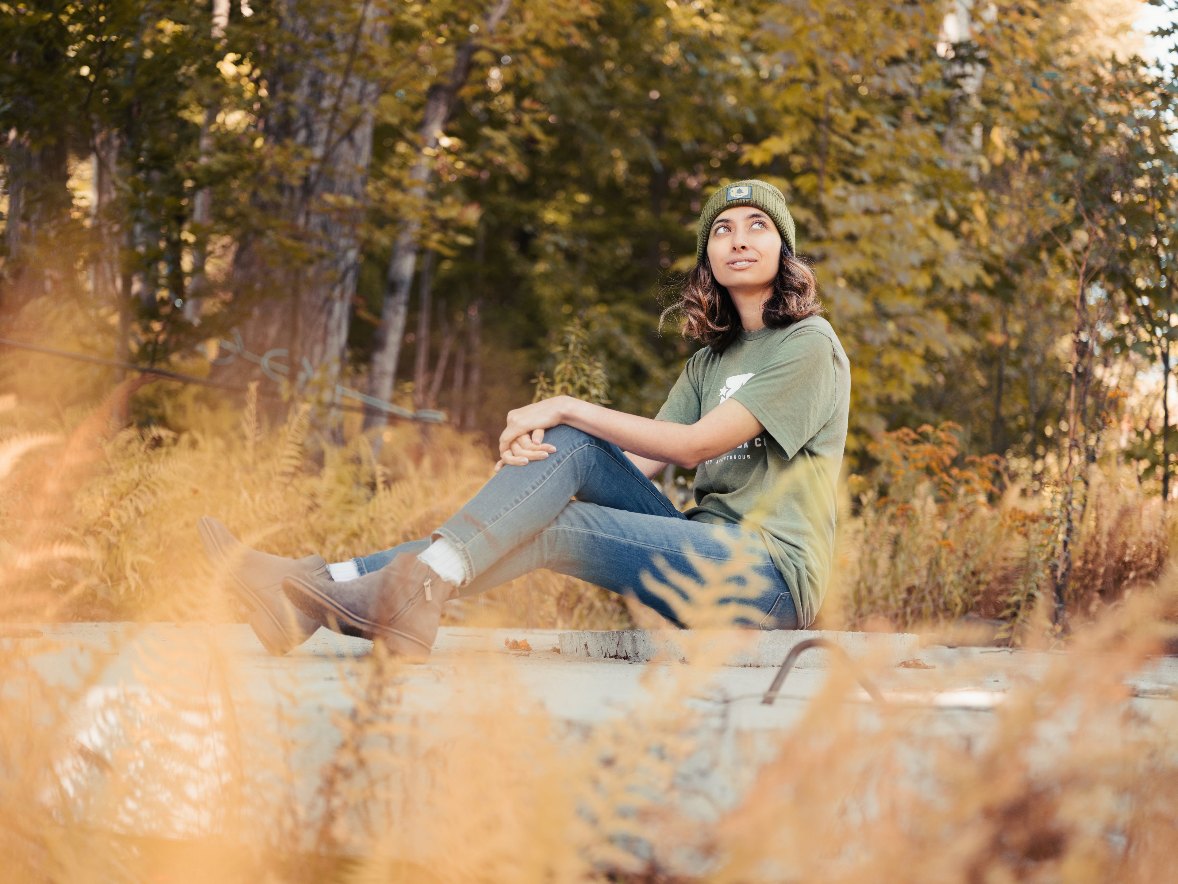 Woman sits in woods wearing Merrimack Co T shirt and beanie 