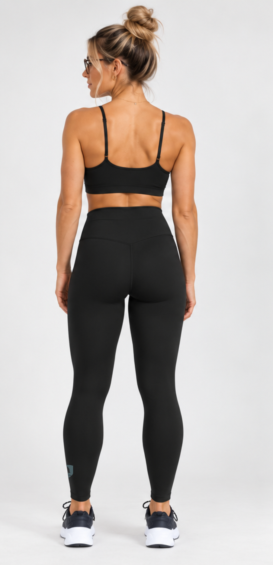 A woman shown from the back wearing a black sports bra and black high-waisted leggings, standing against a light gray studio background.