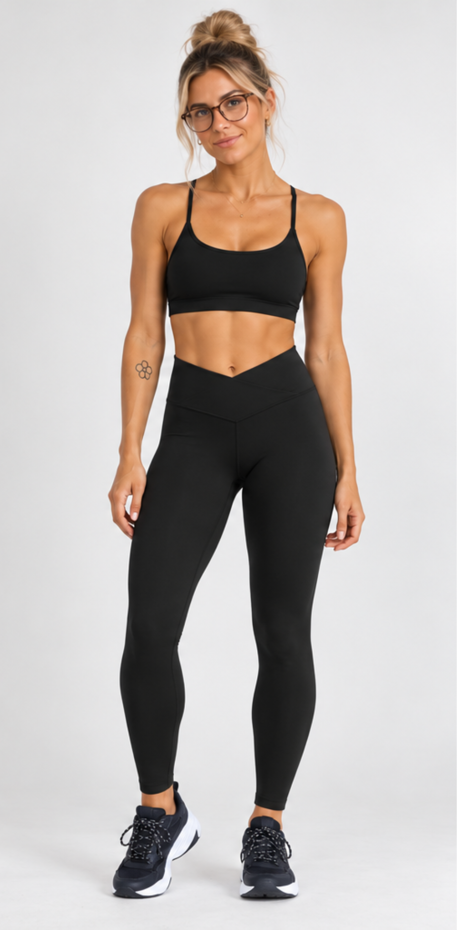 A woman modeling a black sports bra and matching high-waisted black leggings with black athletic sneakers, standing against a light gray studio background.